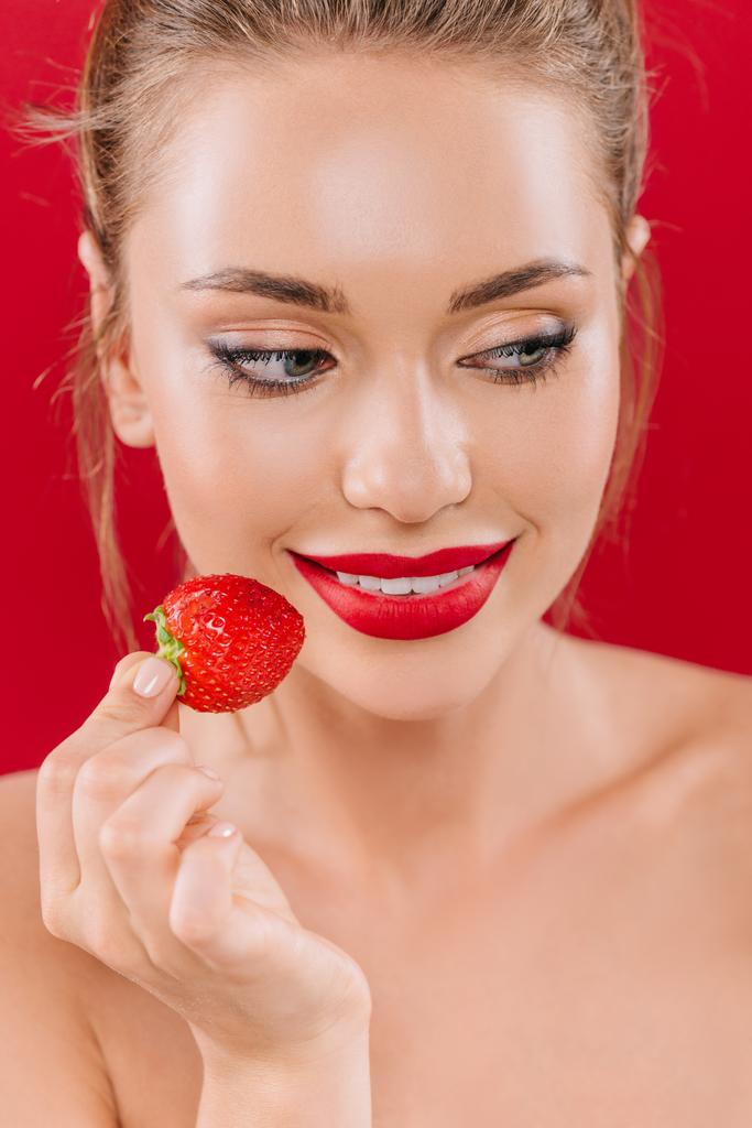 uśmiechnięta naga piękna kobieta z czerwonymi ustami trzymająca truskawkę odizolowaną na czerwono - Zdjęcie, obraz