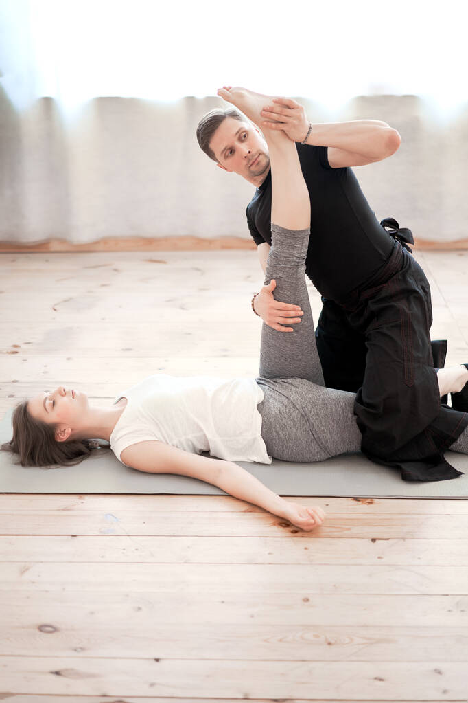 vrouw doen stretching op mat met mannelijke trainer in sportschool tegen achtergrond van venster - Foto, afbeelding