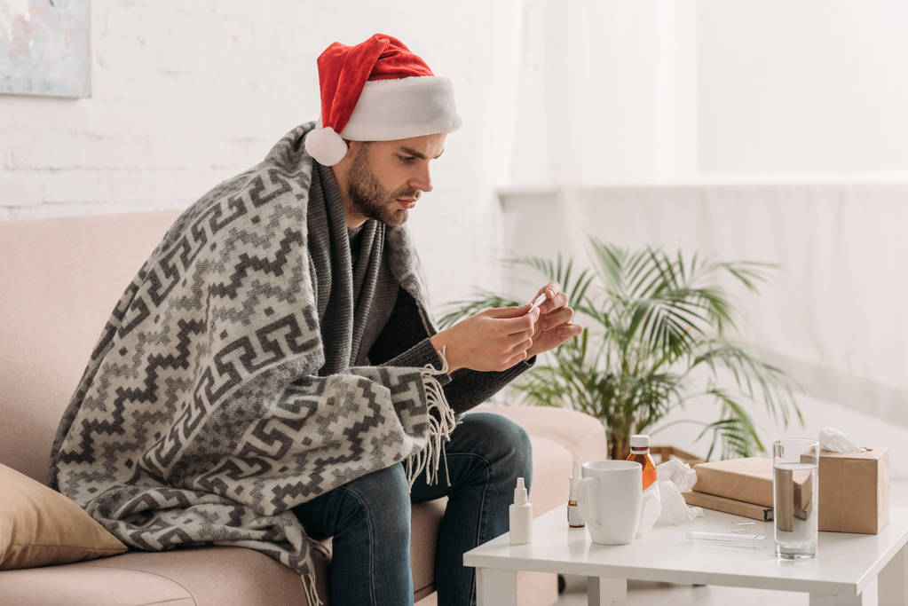 hombre enfermo en sombrero de santa, envuelto en manta, sentado cerca de la mesa con medicinas y sosteniendo el termómetro
 - Foto, imagen