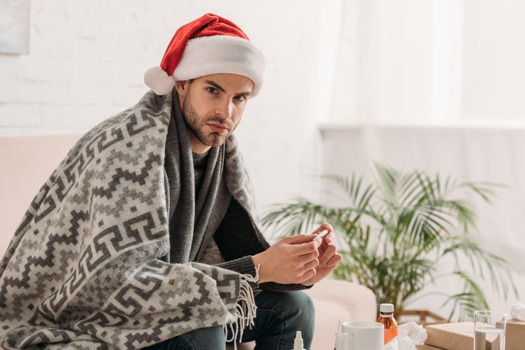 sick man in santa hat, wrapped in blanket, looking at camera while holding thermometer - Photo, Image