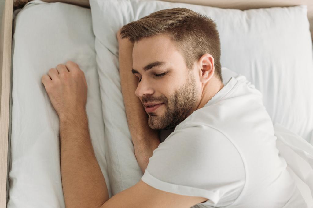 Вид сверху на улыбающегося молодого человека, спящего на белом постельном белье
 - Фото, изображение