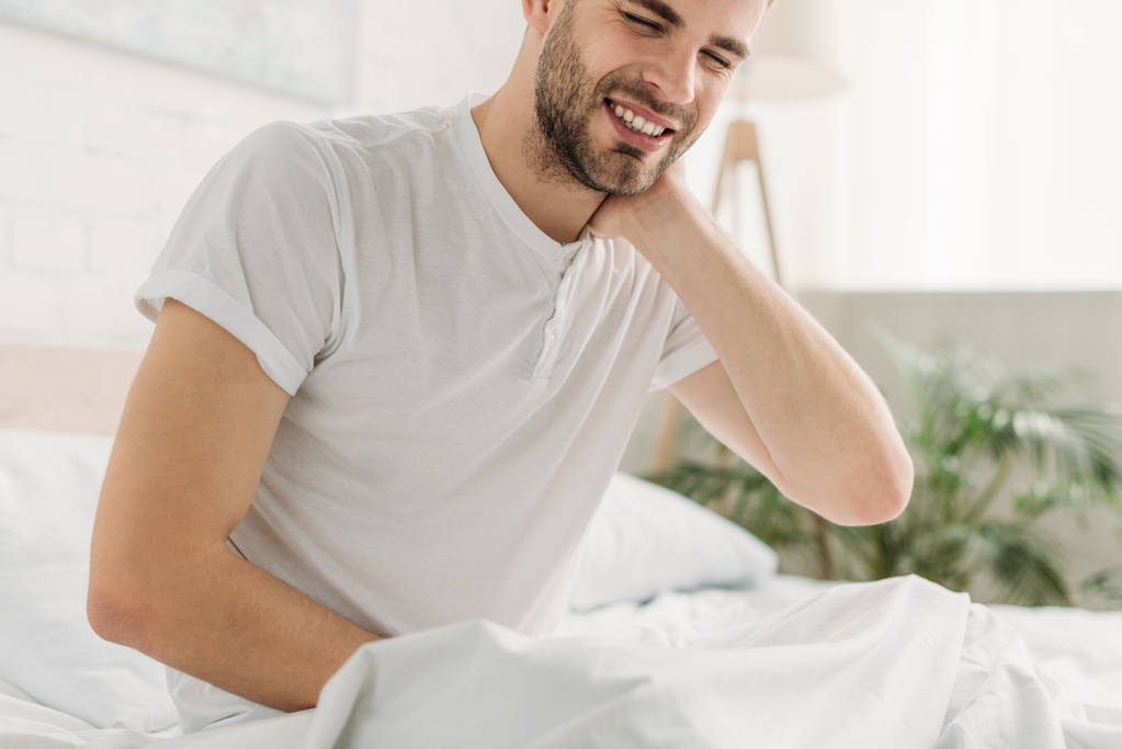 mladý muž sedí na posteli a trpí bolestí krku se zavřenýma očima - Fotografie, Obrázek