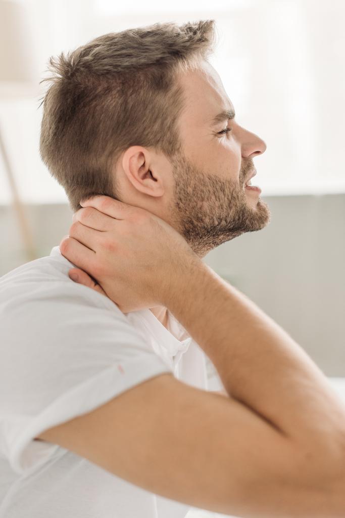 Seitenansicht eines jungen Mannes, der den Hals berührt, während er Schmerzen mit geschlossenen Augen hat - Foto, Bild