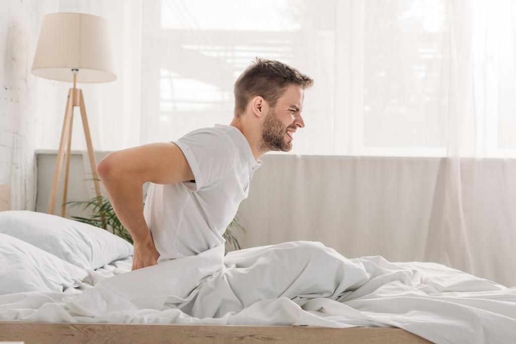 Seitenansicht des verpönten jungen Mannes, der unter Schmerzen im Lendenschurz leidet, während er auf weißem Bettzeug sitzt - Foto, Bild