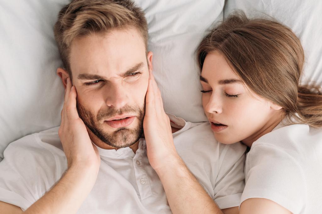 górny widok wyczerpany człowiek zatyka uszy z rąk leżąc w łóżku w pobliżu chrapanie żona - Zdjęcie, obraz