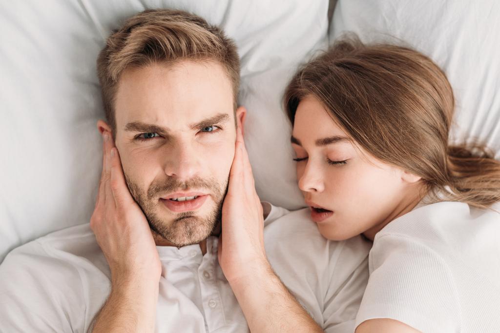 nespokojený muž ucpává uši rukama a dívá se na kameru, zatímco leží v posteli poblíž chrápající manželky - Fotografie, Obrázek