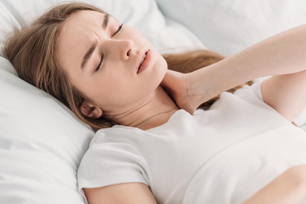 Genç kadın boyun ağrısından muzdaripken gözleri kapalı olarak yatakta yatıyor. - Fotoğraf, Görsel