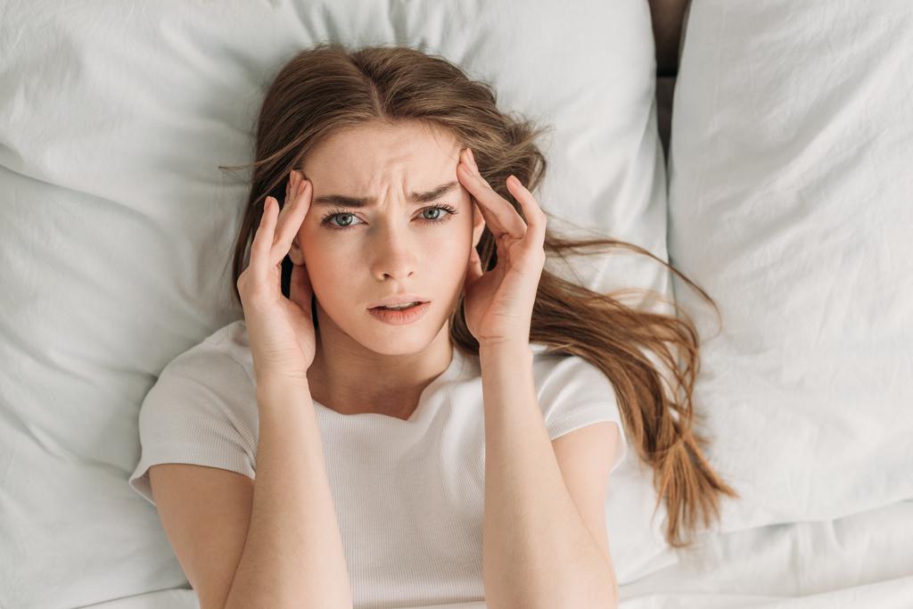 vista superior da jovem mulher olhando para a câmera enquanto deitada na cama e sofrendo de dor de cabeça
 - Foto, Imagem