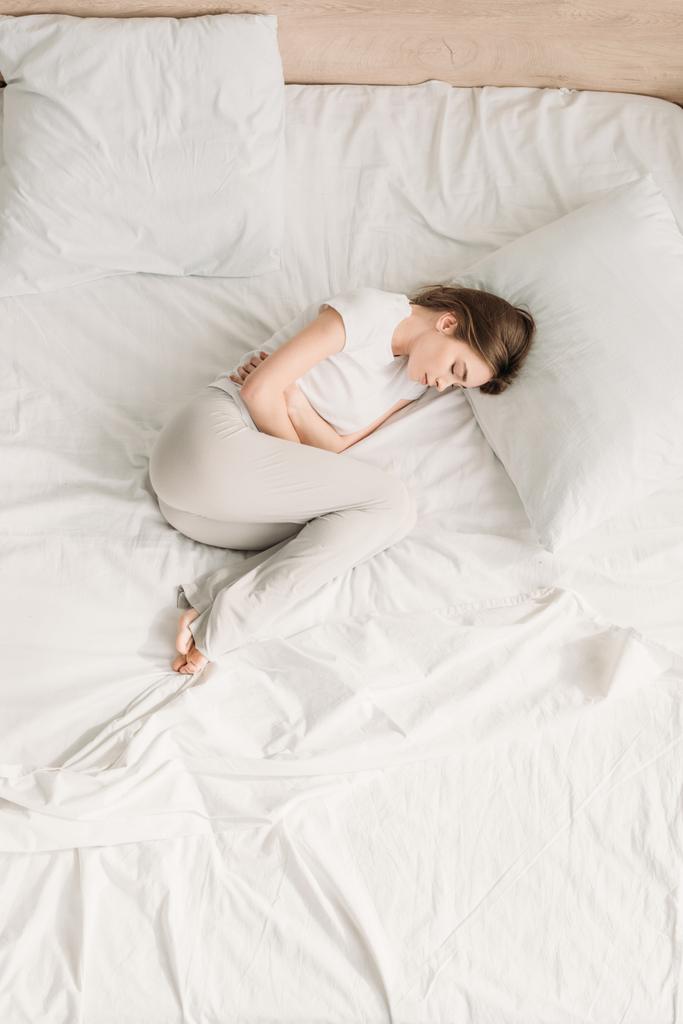 vrchní pohled na dívku ležící v posteli se zavřenýma očima a trpící bolestí břicha - Fotografie, Obrázek