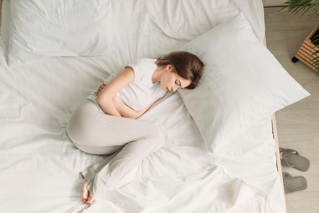 vista superior de la mujer joven acostada en la cama y sufriendo de dolor de estómago
 - Foto, Imagen