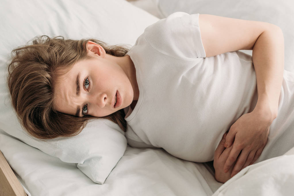 Нахмуренная девушка смотрит в камеру, когда лежит в постели и страдает от боли в животе
 - Фото, изображение
