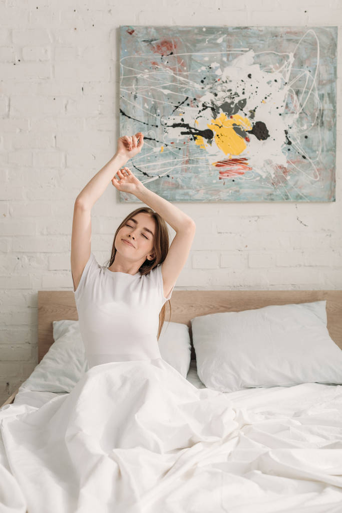 szczęśliwa dziewczyna siedzi w łóżku z zamkniętymi oczami i rozciąga się z podniesionymi rękami - Zdjęcie, obraz