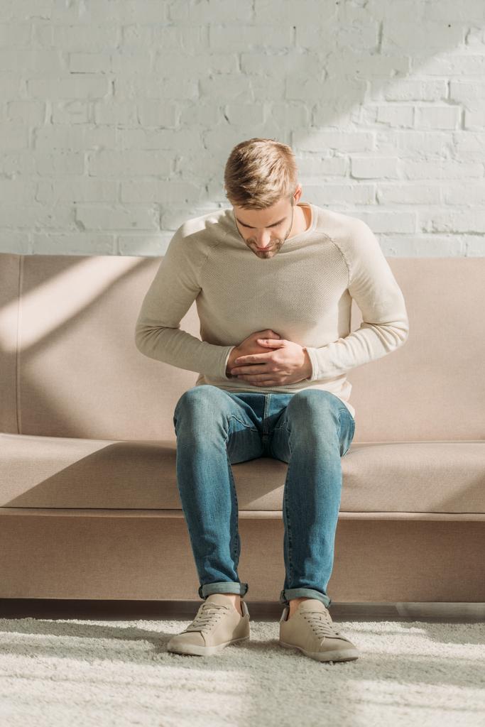 mladý muž sedí na pohovce a dotýká břicha, zatímco trpí bolestí břicha - Fotografie, Obrázek
