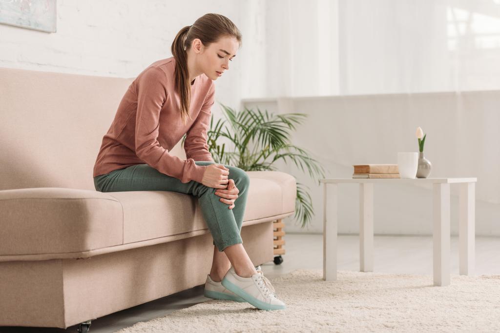 расстроенная девушка сидит на диване и страдает от боли в колене
 - Фото, изображение