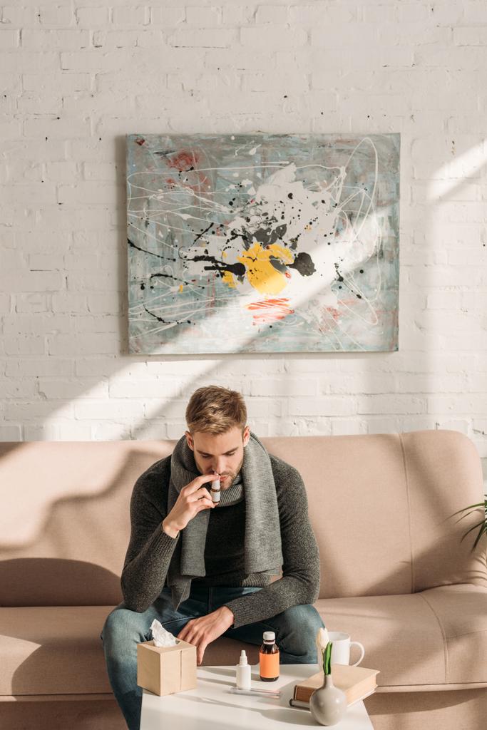 Kranker Mann sitzt mit Medikamenten auf Sofa neben Tisch und benutzt Nasenspray - Foto, Bild