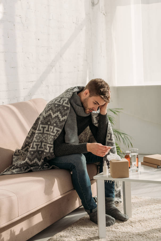 Kranker Mann, in Decke gehüllt, auf Sofa sitzend und auf Thermometer schauend - Foto, Bild