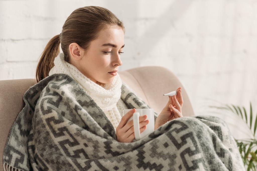 Hastalıklı bir kız, battaniyeye sarılmış, termometreye bakıyor ve elinde bir fincan sıcak içecek tutuyor. - Fotoğraf, Görsel