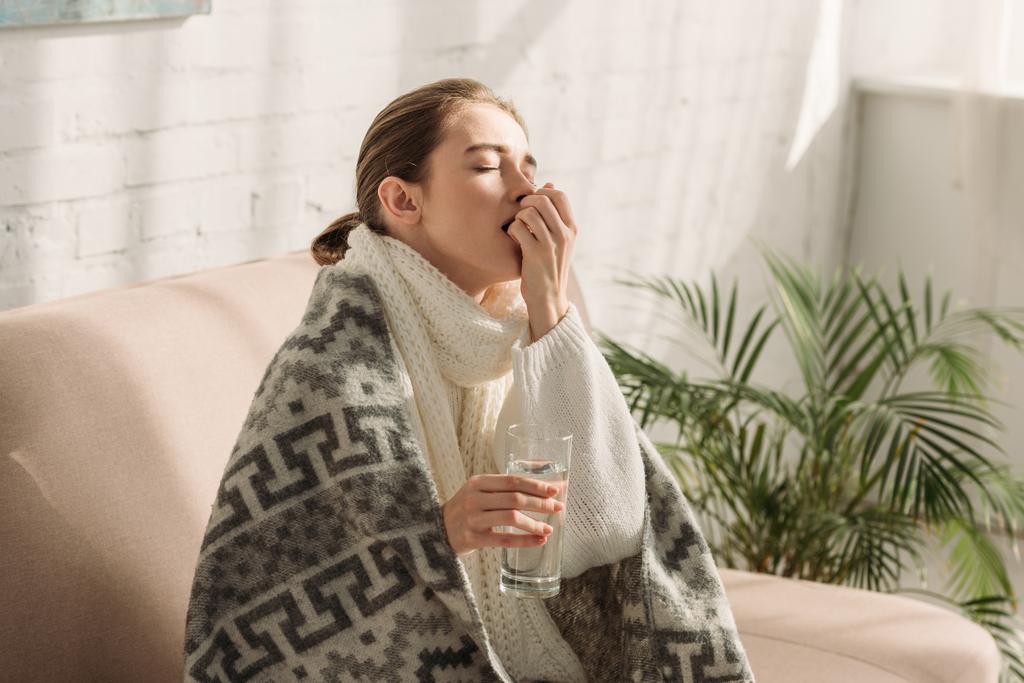 ziek meisje, gewikkeld in een deken, met een glas water en medicijnen - Foto, afbeelding
