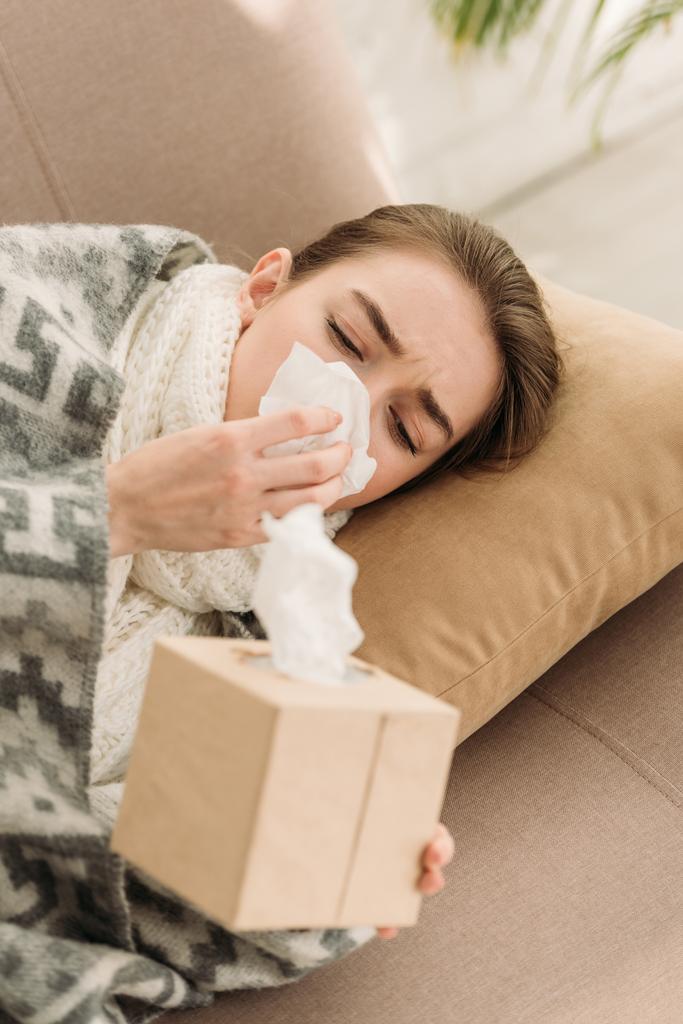 sick girl, covered with blanket, lying on sofa and sneezing in napkin - Photo, Image