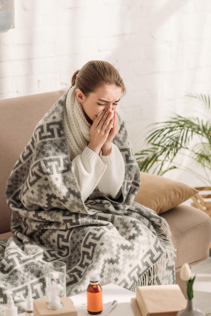 Kranke Frau, in Decke gehüllt, sitzt auf Sofa und niest in Serviette - Foto, Bild