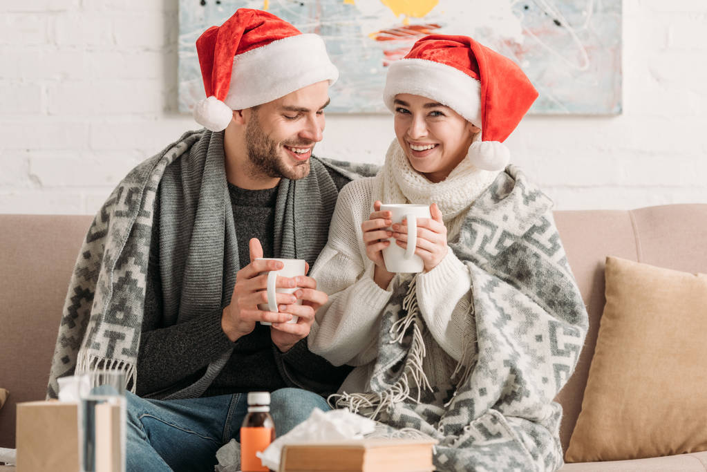 krankes, lächelndes Paar in Weihnachtsmannmützen, mit Decke bedeckt, mit Bechern wärmenden Getränks auf dem Sofa sitzend  - Foto, Bild