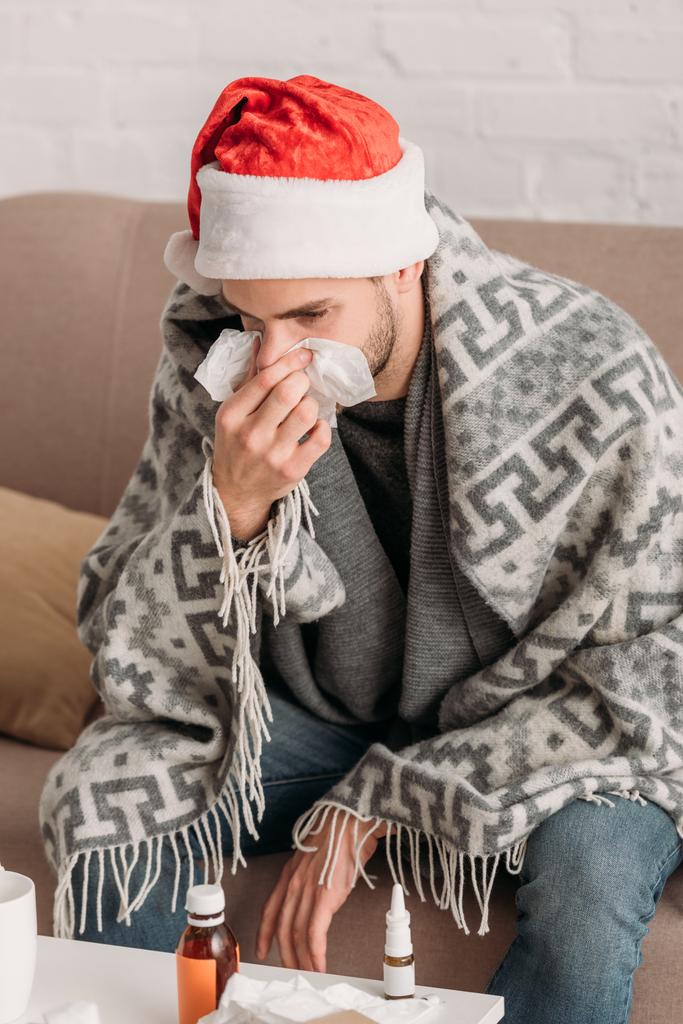 malade assis dans un chapeau de Père Noël, enveloppé dans une couverture, assis dans un canapé et éternuant dans une serviette
 - Photo, image