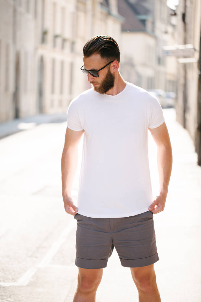 Hipster όμορφο αρσενικό μοντέλο με γενειάδα φορώντας λευκό κενό t-shirt και ένα καπέλο του μπέιζμπολ με χώρο για το λογότυπο ή το σχέδιό σας σε casual αστικό στυλ - Φωτογραφία, εικόνα
