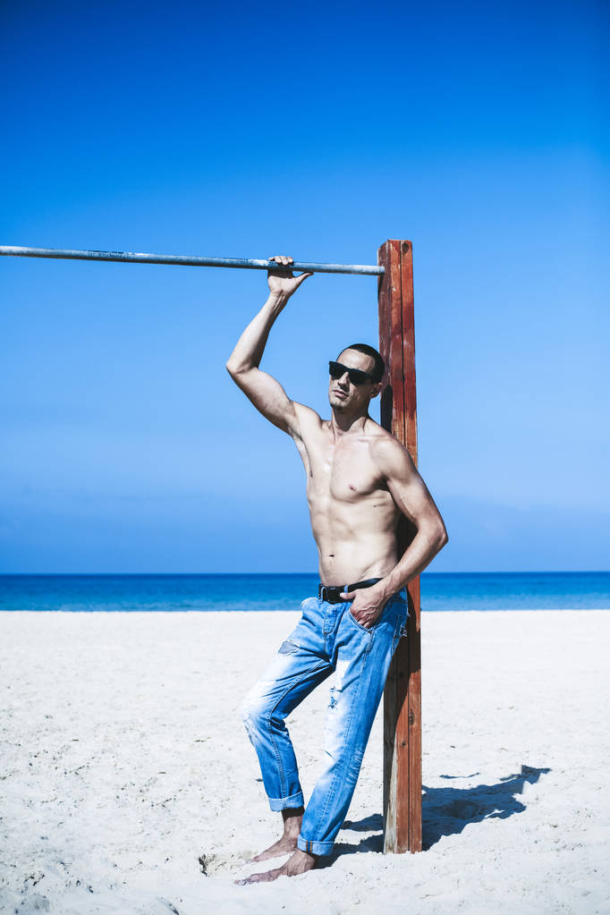 jonge gespierde man rustend en poserend op het strand. Zonnebrillen dragen - Foto, afbeelding