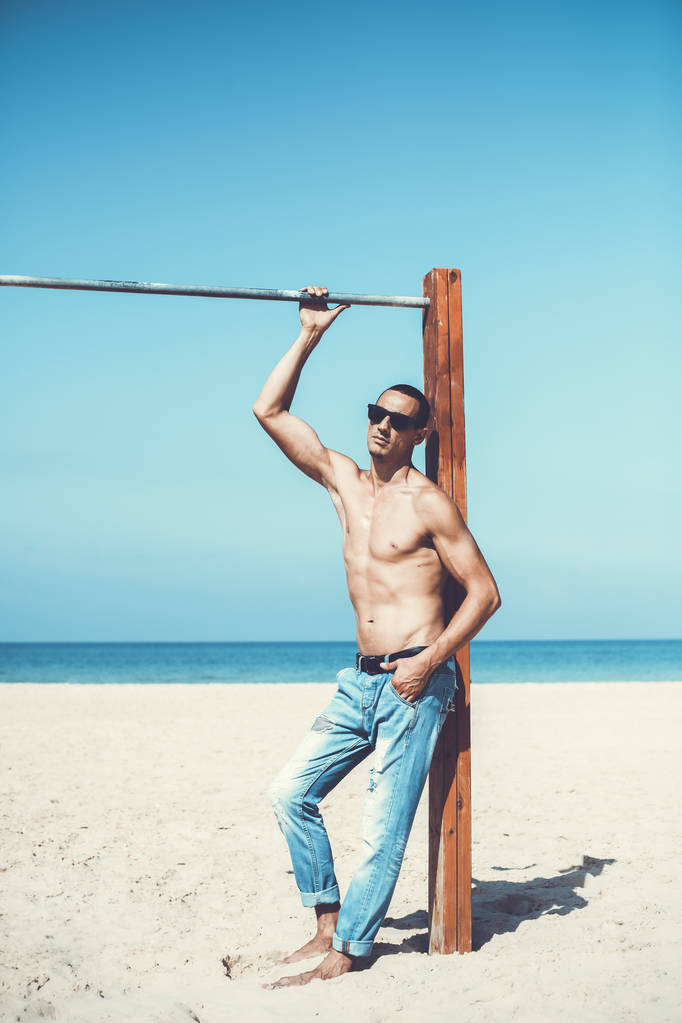 nuori lihaksikas mies lepää ja poseeraa rannalla. Aurinkolasien käyttö
 - Valokuva, kuva