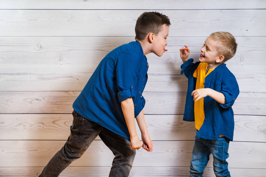Two boys in casual clothes in white wooden background showing tongues to each other. Two brothers playing. Elder and smaller brothers. Happy childhood concept. Frienship. - Valokuva, kuva