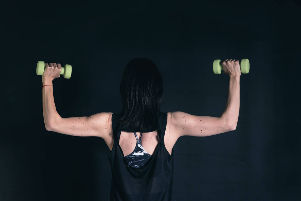 Kolları için egzersiz yapan atletik bir kadın. Siyah arkaplanda dambıllarla çalışmak - Fotoğraf, Görsel