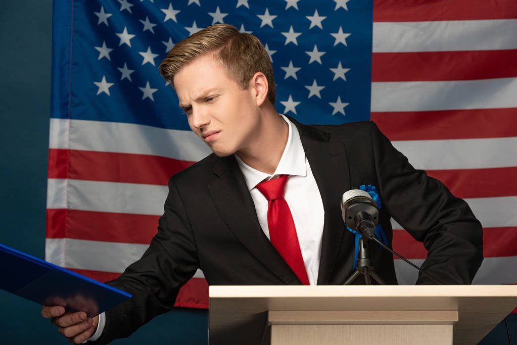 ontevreden man kijkt naar klembord op tribune op Amerikaanse vlag achtergrond - Foto, afbeelding