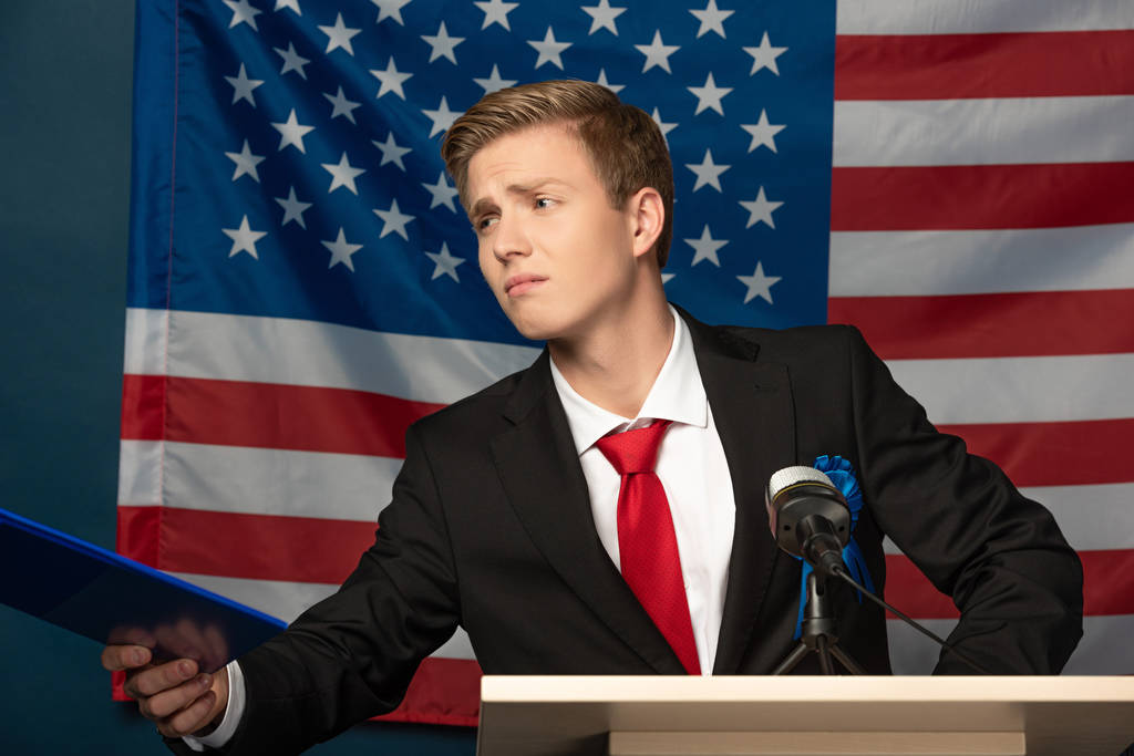 Unzufriedener Mann hält Klemmbrett auf Tribüne vor Hintergrund amerikanischer Flagge - Foto, Bild