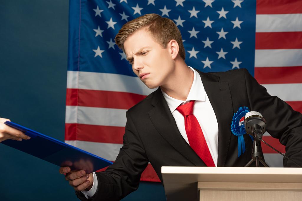 displeased man holding clipboard on tribune on american flag background - 写真・画像