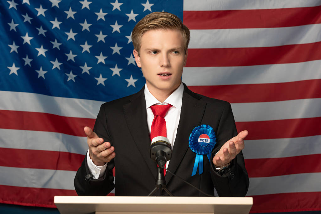 Emotionaler Mann auf der Tribüne während einer Rede vor dem Hintergrund der amerikanischen Flagge - Foto, Bild