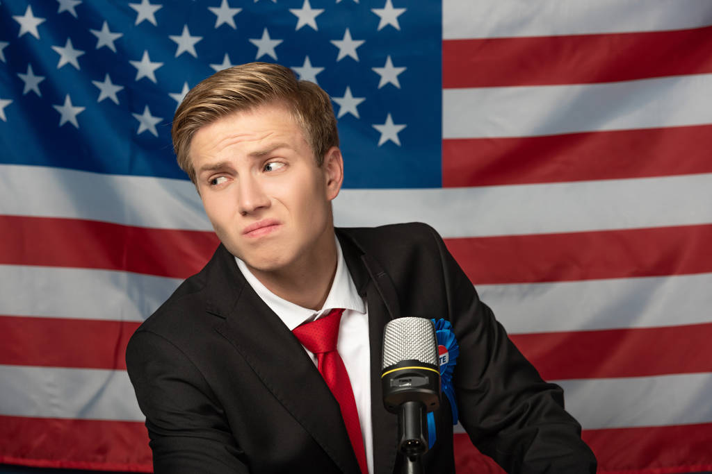 смущенный человек на трибуне на фоне американского флага
 - Фото, изображение