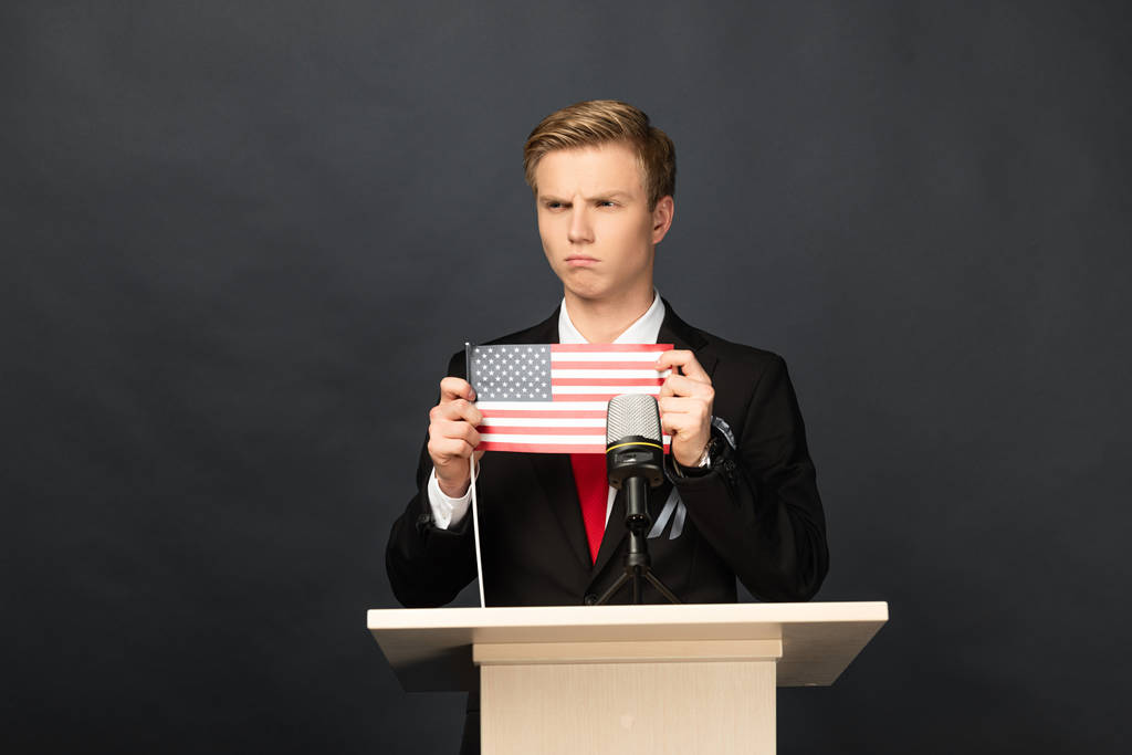 сердитый эмоциональный человек на трибуне с американским флагом на черном фоне
 - Фото, изображение