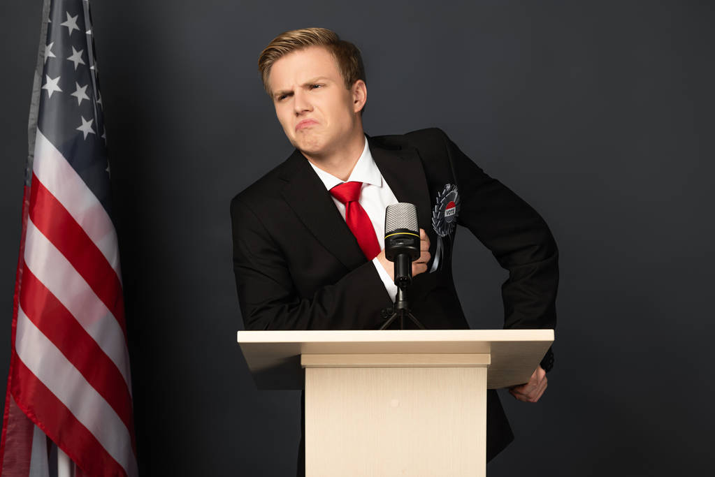 Unzufriedener emotionaler Mann auf der Tribüne mit amerikanischer Flagge auf schwarzem Hintergrund - Foto, Bild