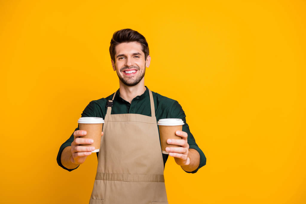 Portrait of positive cheerful guy worker in coffee bar shop hold paper card cup offer to his client mug with hot cappuccino wear apron green shirt isolated over yellow color background - Photo, Image
