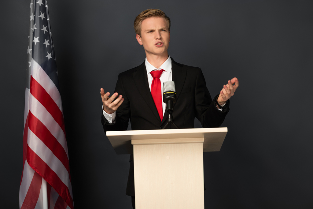 emotionele man spreekt op tribune met Amerikaanse vlag op zwarte achtergrond - Foto, afbeelding