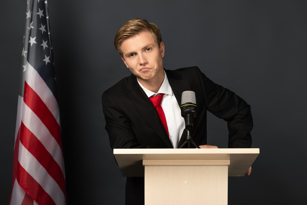 ontevreden emotionele man op tribune met Amerikaanse vlag op zwarte achtergrond - Foto, afbeelding