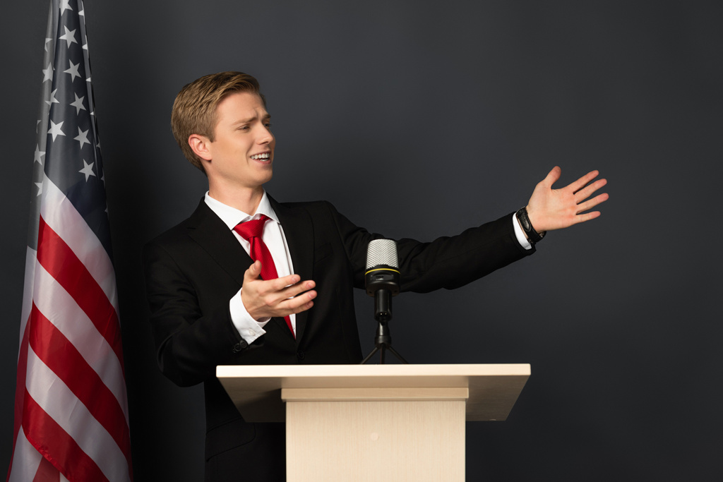 hombre emocional hablando y haciendo gestos en tribuna con bandera americana sobre fondo negro
 - Foto, imagen