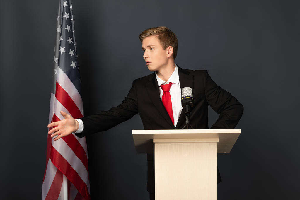 Emotionaler Mann spricht und gestikuliert auf Tribüne mit amerikanischer Flagge auf schwarzem Hintergrund - Foto, Bild