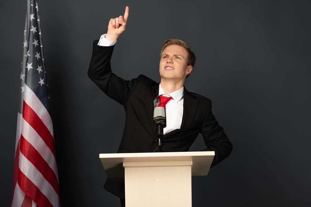 emotivní muž ukazuje prstem nahoru na tribuna s americkou vlajkou na černém pozadí - Fotografie, Obrázek