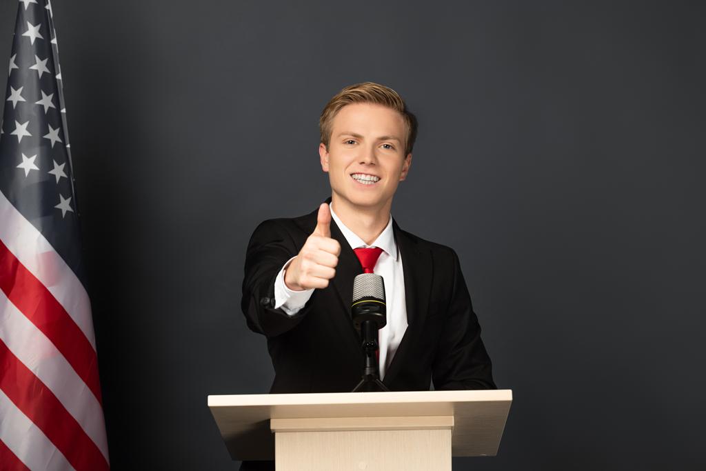 sorrindo homem mostrando polegar em tribuno com bandeira americana em fundo preto
 - Foto, Imagem