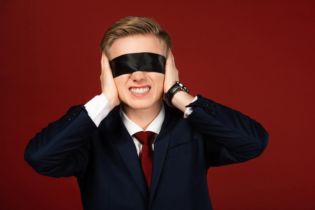 man met blinddoek op ogen oren bedekkend met handen op rode achtergrond - Foto, afbeelding