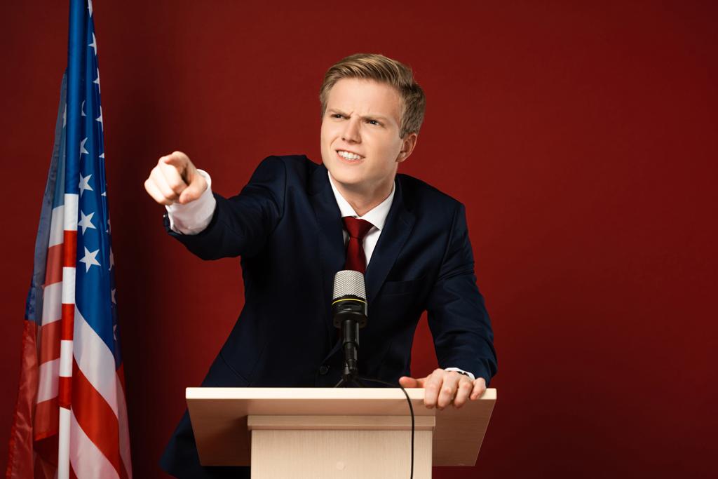 συναισθηματικός άνθρωπος που δείχνει με το δάχτυλο σε tribune κοντά αμερικανική σημαία σε κόκκινο φόντο - Φωτογραφία, εικόνα