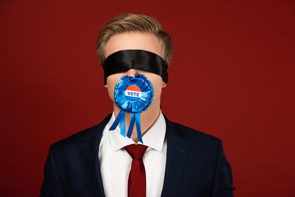 homem imitando com venda nos olhos e crachá com letras de voto na boca no fundo vermelho
 - Foto, Imagem