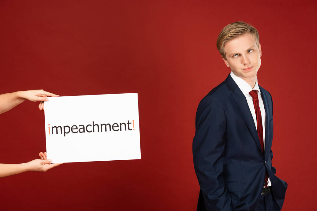 sceptyczny mężczyzna grymaszący w pobliżu białej karty z napisem impeachment na czerwonym tle - Zdjęcie, obraz