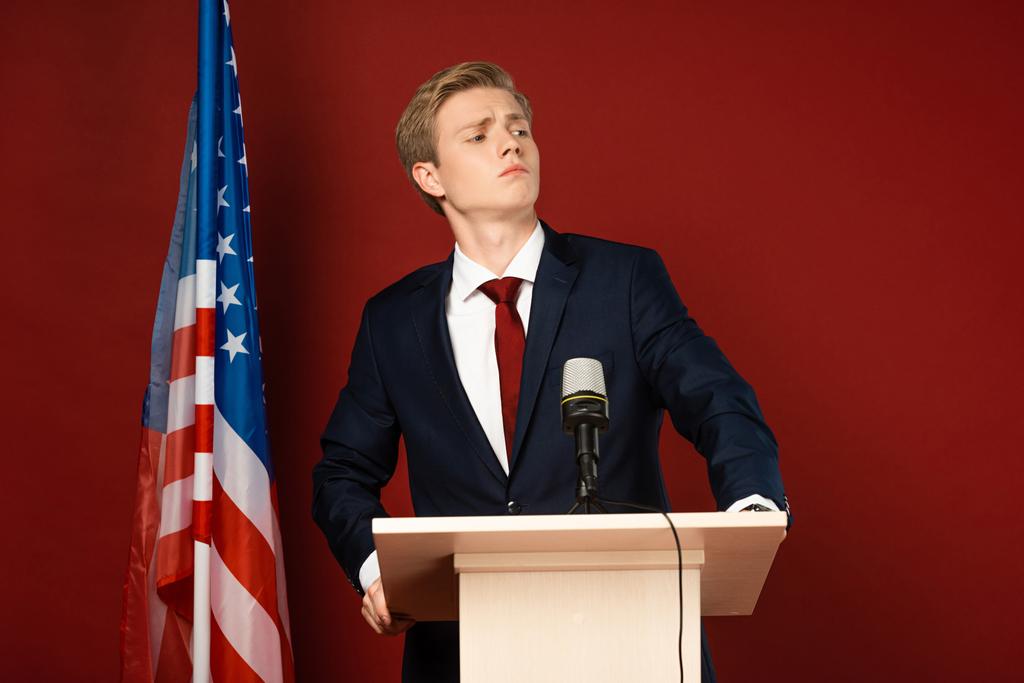 ernster Mann steht auf der Tribüne neben der amerikanischen Flagge auf rotem Hintergrund - Foto, Bild